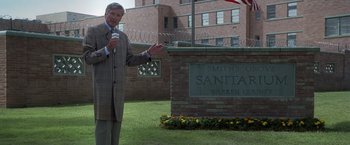 Movie still from “Halloween” (2007), directed by Rob Zombie – A man in a brown suit is holding a microphone in front of a sign; Wide shot, High angle