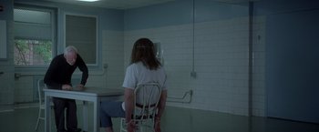 Movie still from “Halloween” (2007), directed by Rob Zombie – A man sitting on a chair in a room; Wide shot, Over the shoulder angle