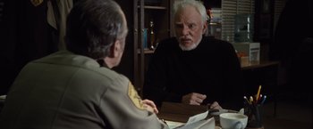 Movie still from “Halloween” (2007), directed by Rob Zombie – A man sitting at a table talking to another man in front of a book; Medium shot, Over the shoulder angle