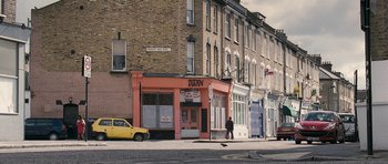 Movie still from “Happy-Go-Lucky” (2008), directed by Mike Leigh – A person walking down the street in front of a building; Extreme Wide shot, High angle