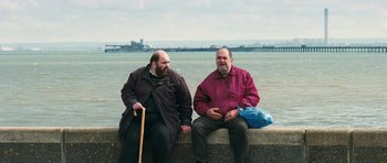 Movie still from “Happy-Go-Lucky” (2008), directed by Mike Leigh – Two men sitting next to a body of water; Medium shot, Low angle