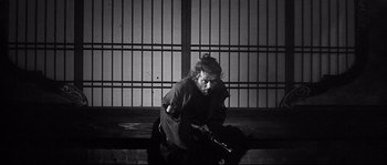 Movie still from “Harakiri” (1962), directed by Masaki Kobayashi – An older man sitting on a bench with a gun in his hand; Medium shot, Over the shoulder angle