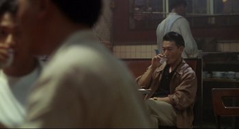 Movie still from “Hard Boiled” (1992), directed by John Woo – A man sitting at a table with a drink in his hand; Medium shot, Over the shoulder angle
