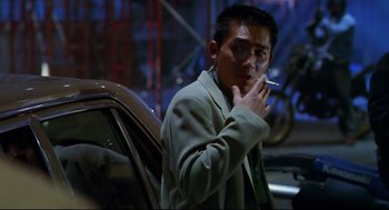 Movie still from “Hard Boiled” (1992), directed by John Woo – A man standing next to a parked car smoking a cigarette; Medium shot, Low angle