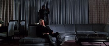 Movie still from “Hard Eight” (1996), directed by Paul Thomas Anderson – A man sitting on top of a couch in a living room; Wide shot, Over the shoulder angle