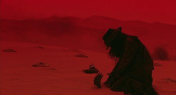 Movie still from “Hardware” (1990), directed by Richard Stanley – A man in a hat and a long black coat kneeling on the ground; Wide shot, High angle