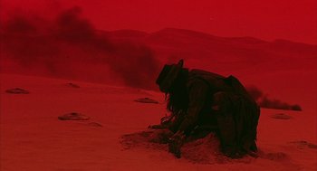 Movie still from “Hardware” (1990), directed by Richard Stanley – A man in a hat is kneeling in the sand; Wide shot, High angle