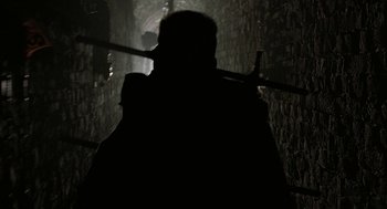 Movie still from “Hardware” (1990), directed by Richard Stanley – A person is holding a sword in a dark room; Wide shot, Over the shoulder angle