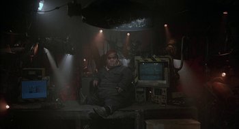 Movie still from “Hardware” (1990), directed by Richard Stanley – A man sitting on a chair next to a television; Wide shot, High angle