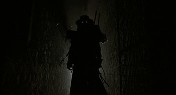 Movie still from “Hardware” (1990), directed by Richard Stanley – A man in a trench coat standing in the dark; Wide shot, Low angle