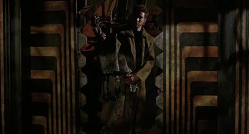 Movie still from “Hardware” (1990), directed by Richard Stanley – A man holding a walking stick standing in front of a painting; Wide shot, Low angle