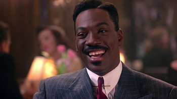 Movie still from “Harlem Nights” (1989), directed by Eddie Murphy – A man wearing a suit and tie smiling for the camera; Close Up shot, Over the shoulder angle