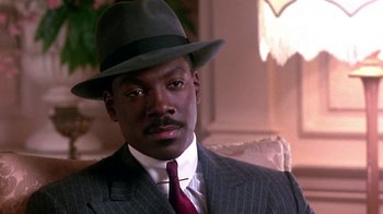 Movie still from “Harlem Nights” (1989), directed by Eddie Murphy – A man wearing a suit and a hat is sitting in a chair; Close Up shot, Over the shoulder angle